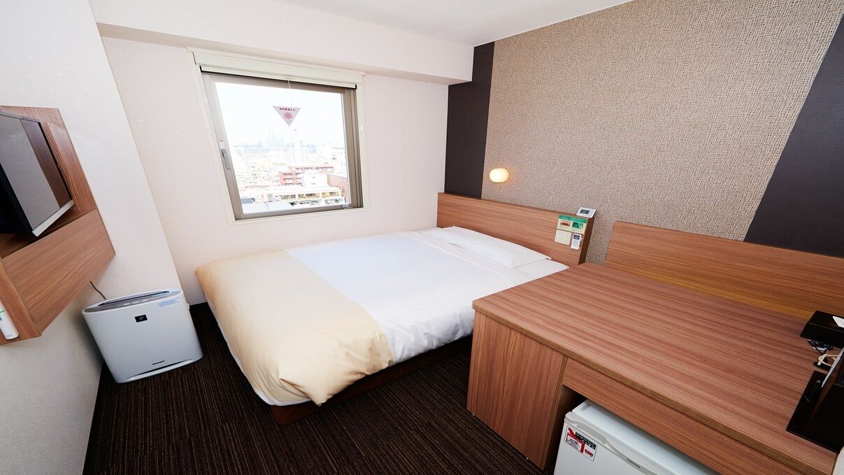 Ein Hotelzimmer mit einem Bett, einem Schreibtisch, einem kleinen Heizgerät, einem Fenster und einem an der Wand befestigten Fernseher.