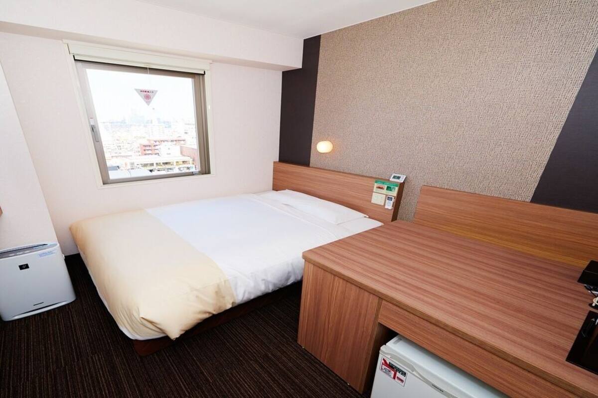 Une chambre d’hôtel équipée d’un lit, d’un bureau, d’un petit radiateur, d’une fenêtre et d’un téléviseur fixé au mur.