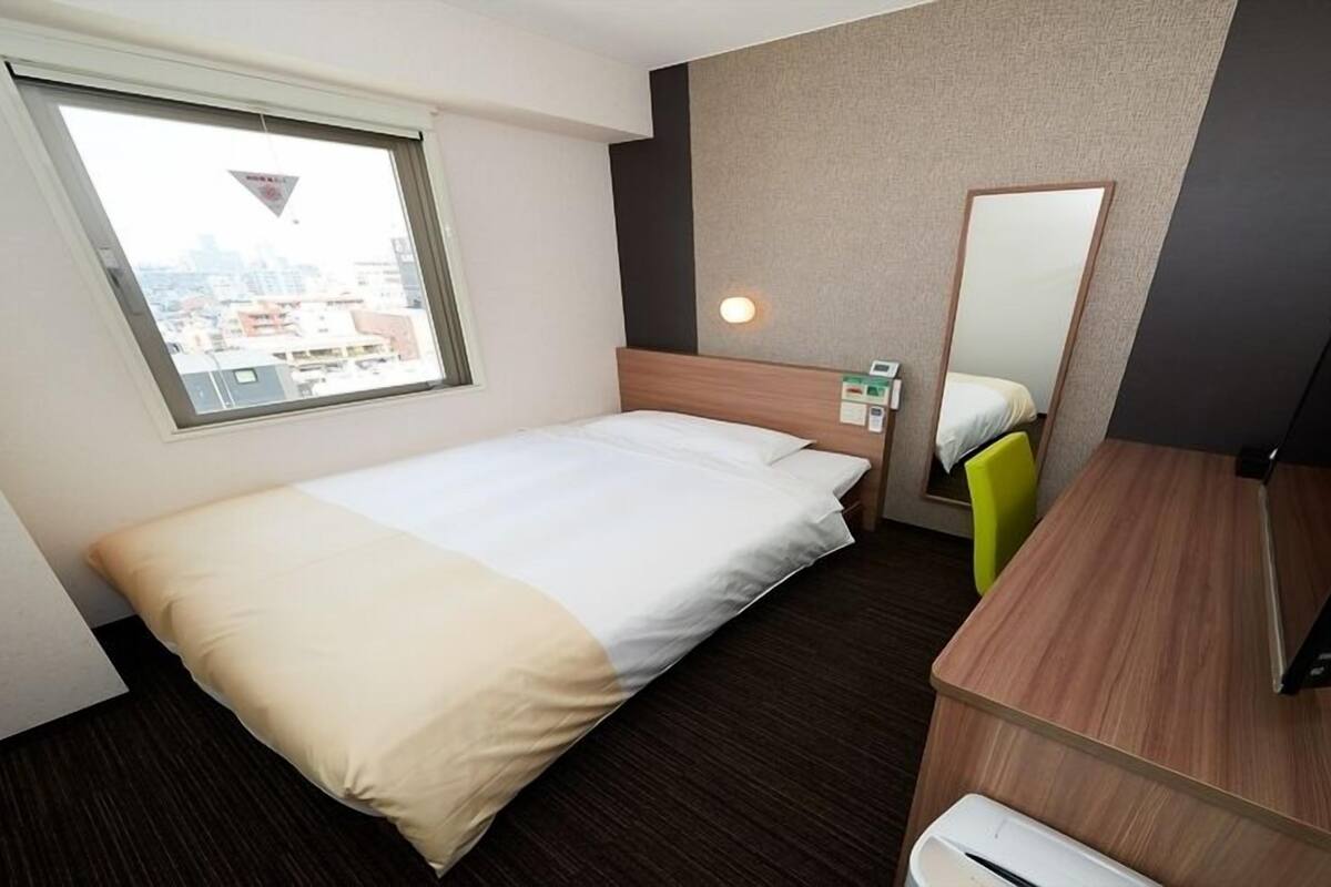 Une chambre d’hôtel avec un lit, un bureau, une chaise, un miroir et une fenêtre.