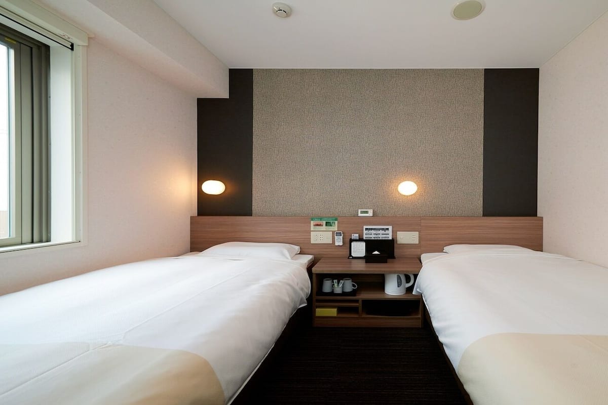 Une chambre d’hôtel avec deux lits simples, une tête de lit en bois, un petit bureau avec un téléphone et une machine à café, ainsi qu’un luminaire fixé au mur.
