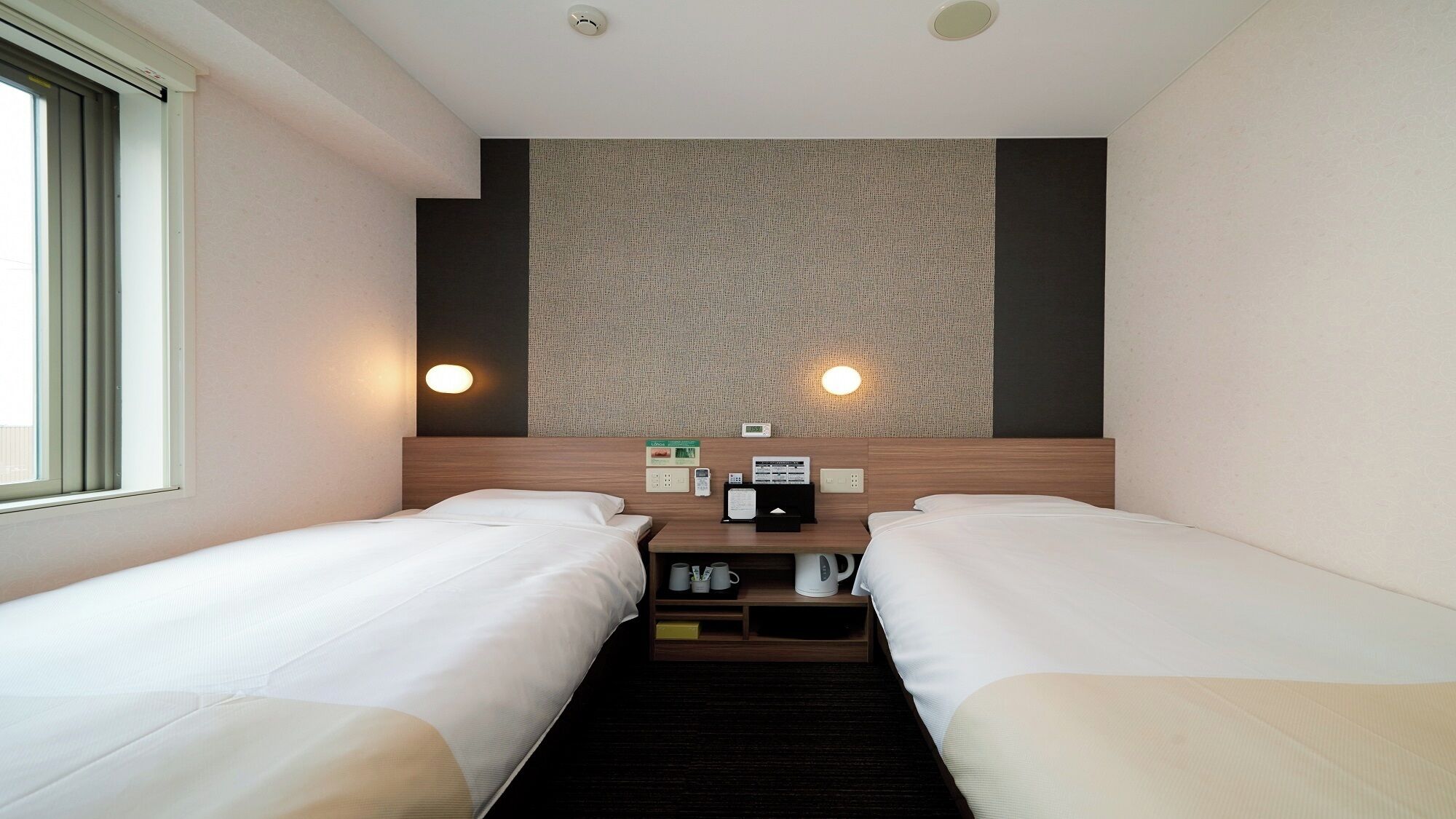 Ein Hotelzimmer mit zwei Betten, einem Kopfteil mit strukturierter Wand, Nachttischlampen, einer Bedienkonsole und einem Fenster.