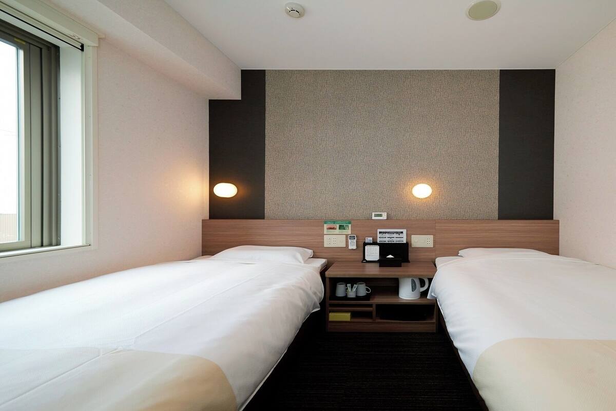 Une chambre d’hôtel avec deux lits, une tête de lit agrémentée d’un mur texturé, des lampes de chevet, un panneau de commande et une fenêtre.