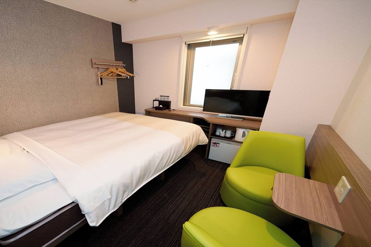 Une chambre d’hôtel avec un lit, un bureau avec une télévision, un petit réfrigérateur et une chaise verte.