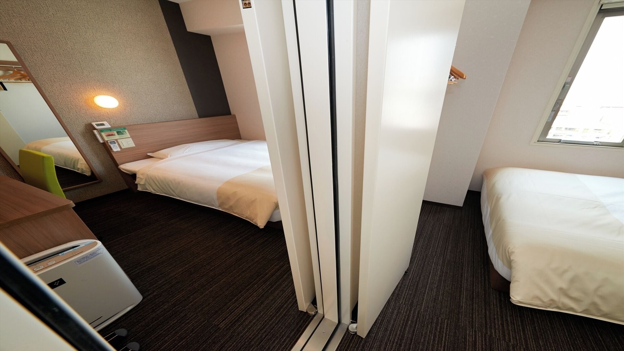Ein kleines Hotelzimmer mit zwei Betten, einem Schreibtisch, einem Stuhl, einem kleinen Tisch, einem Wandheizkörper und einem Fenster.