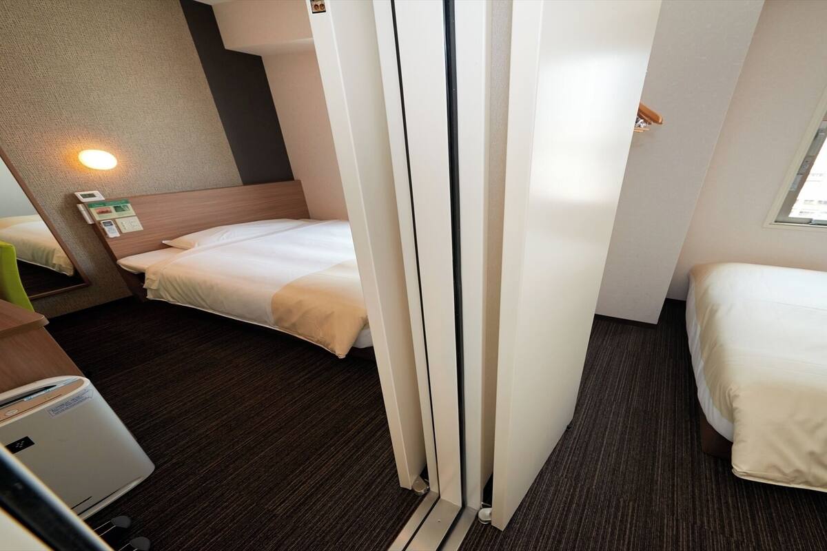 Une chambre d’hôtel de taille réduite, comprenant deux lits, un bureau, une chaise, une petite table, un radiateur fixé au mur et une fenêtre.