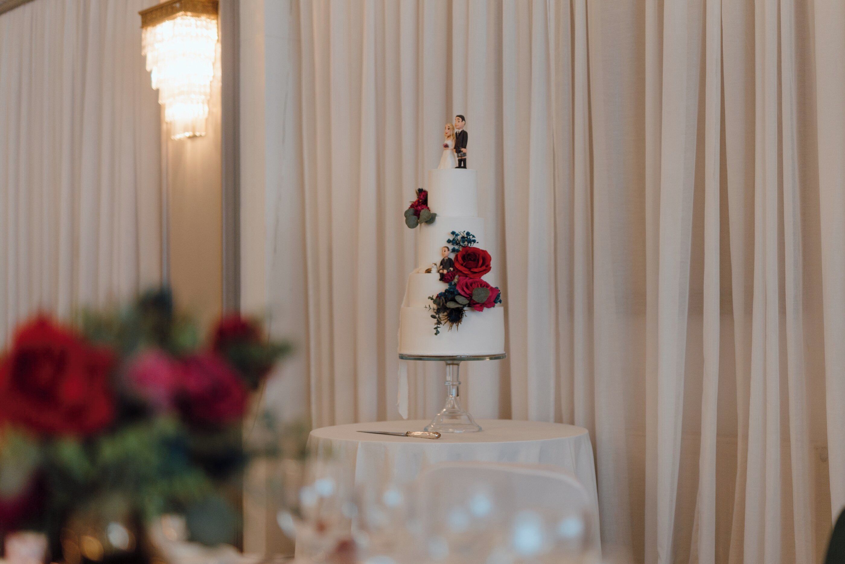 Un pastel de bodas de varios niveles con decoraciones florales y una figurita de una pareja en la cima, sobre una mesa con mantel blanco.