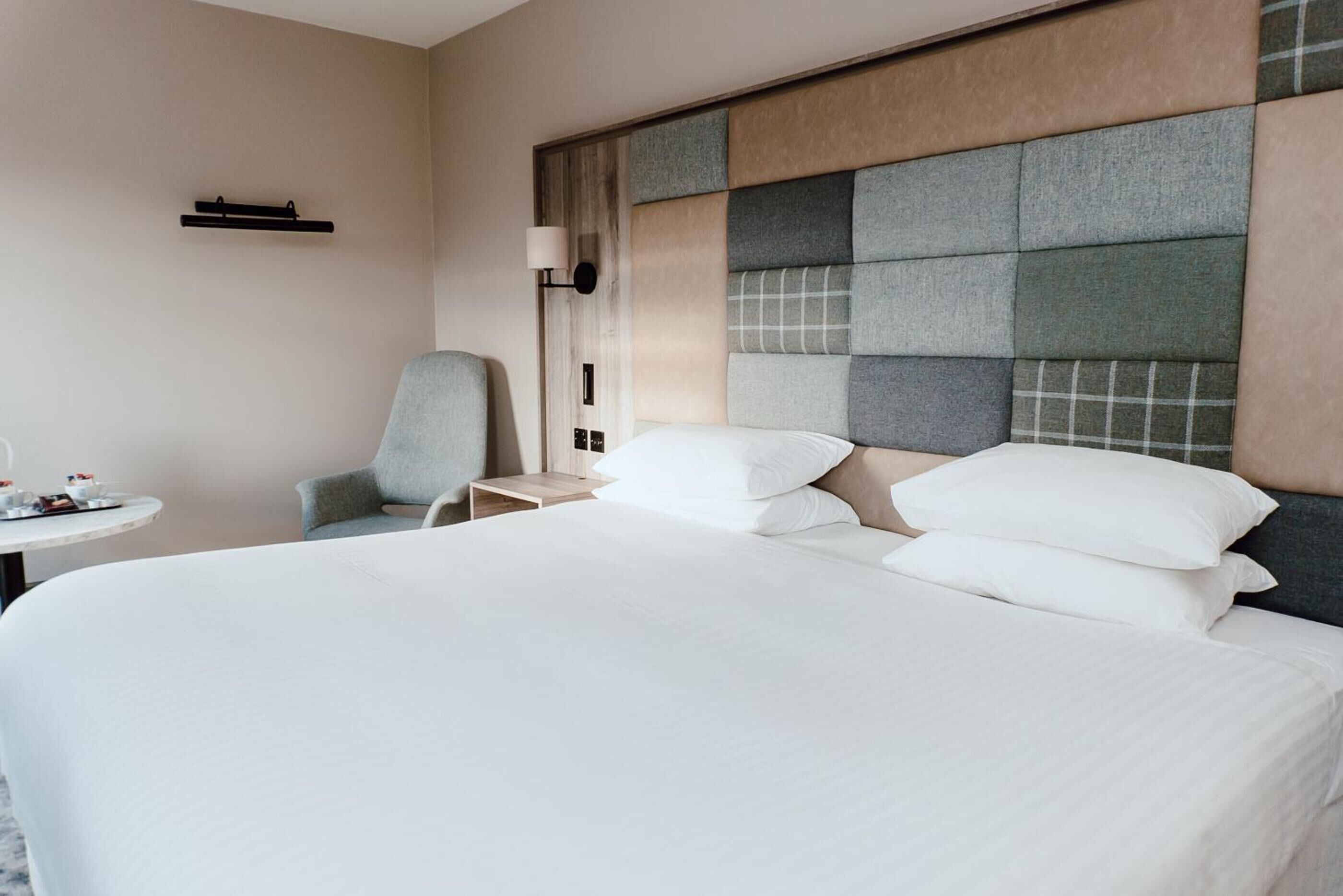 A hotel room with a large bed, a chair, a small table with items, and a wall-mounted lamp.
