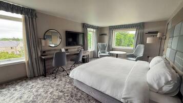 A hotel room with a large bed, two armchairs, a desk, a TV, and a window with a view of greenery.