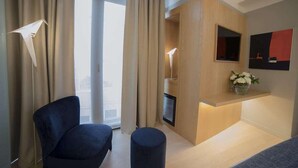 A modern hotel room with a large window, a dark blue chair, a small ottoman, a wooden accent wall, a mounted TV, and a small shelf with flowers. - Galata's Hotel Istanbul (Istanbul)