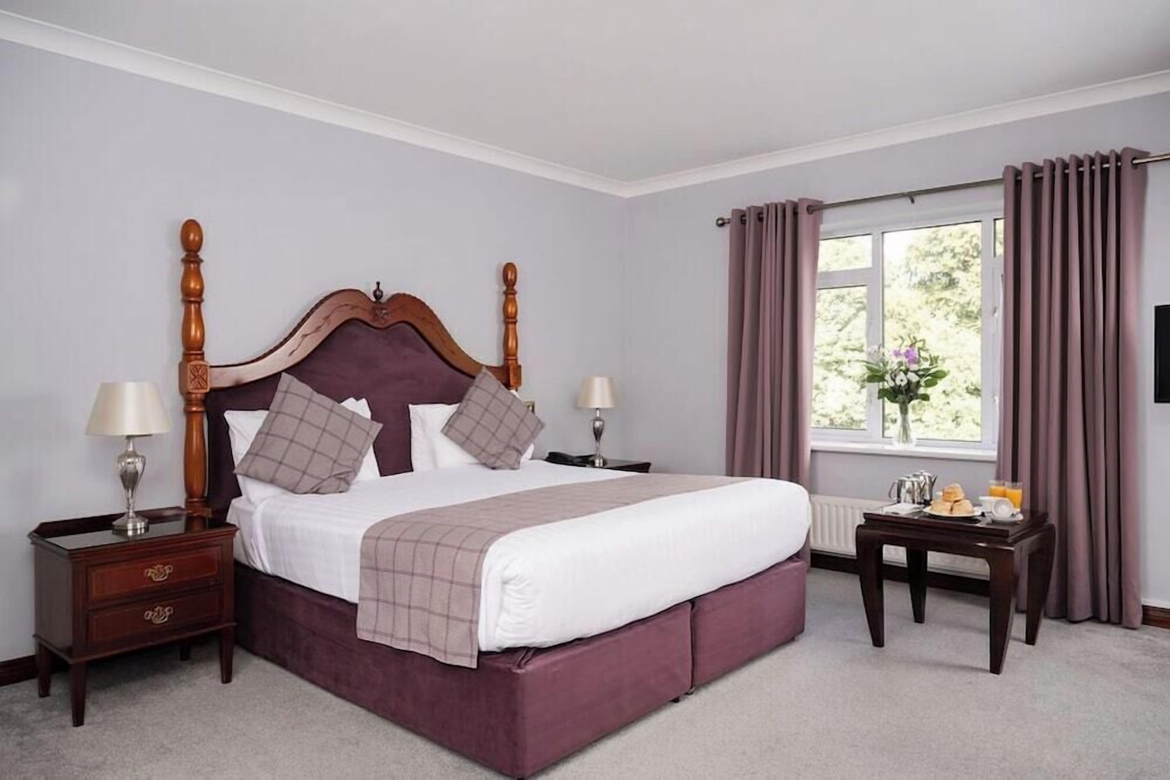 A neatly made bed with a wooden headboard, two bedside tables with lamps, a window with curtains, and a small table with a vase and items on it.