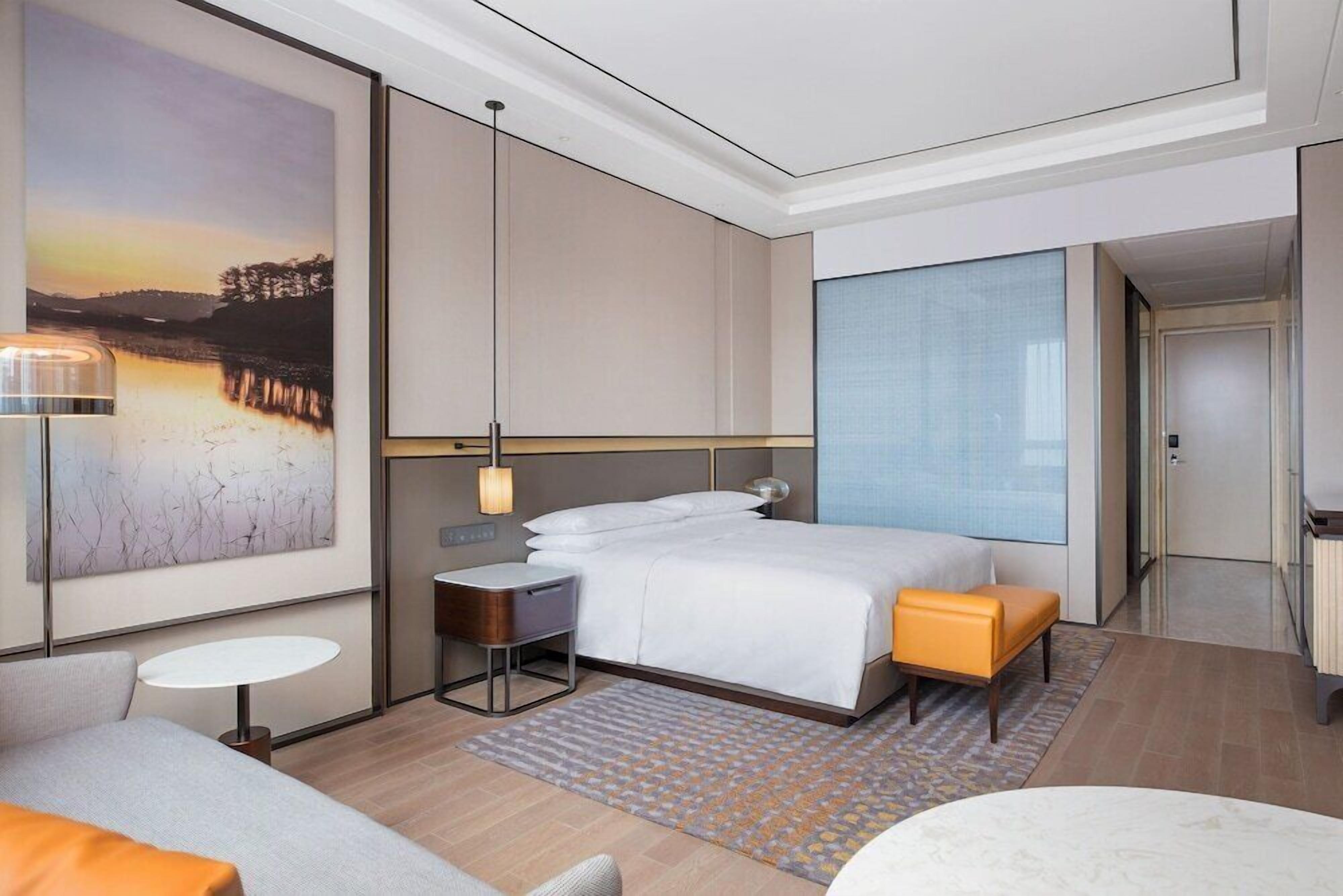 A modern hotel room with a large bed, a small desk, a seating area, and a large window.