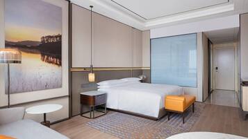 A modern hotel room with a large bed, a small desk, a seating area, and a large window.