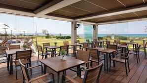 A covered outdoor dining area with wooden tables and chairs, large windows offering a view of the ocean and greenery.