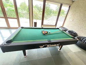A pool table with balls and a cue stick, located in a room with large windows and a brick wall. - Lebua Corbett (Salt)