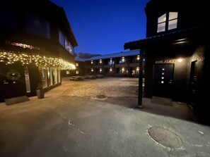 Entrada de hotel com área externa coberta, balcão de recepção e um edifício com letreiros iluminados.