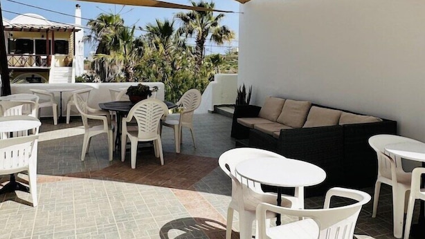 An outdoor patio area with white plastic furniture, including tables and chairs, a black sofa, and a large beige canopy providing shade. - Arlene (Santorini)