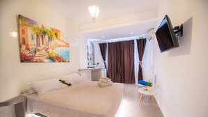 A modern hotel room with a large bed, a flat-screen TV mounted on the wall, and a painting of a coastal scene. - Arlene (Santorini)