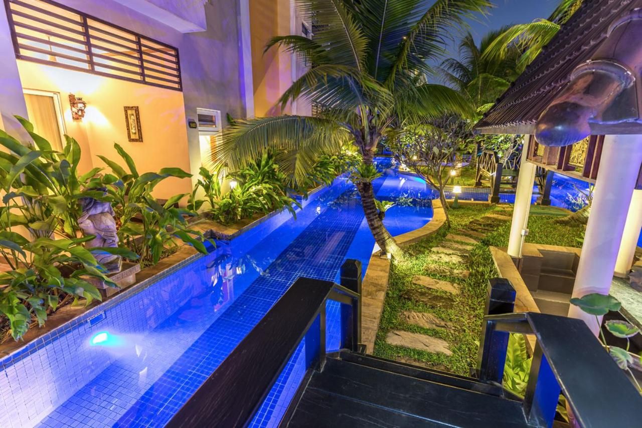 A pool area with a blue-tiled pathway, palm trees, and a building with a tiled roof.