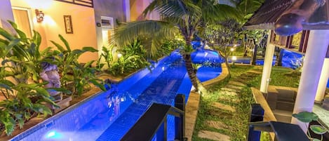 A pool area with a blue-tiled pathway, palm trees, and a building with a tiled roof.