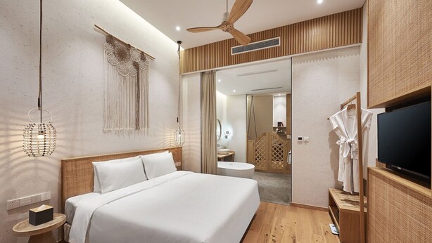 Una habitación de hotel moderna con una cama grande, ventilador de techo, suelo de madera, televisor y baño visible a través de una puerta abierta.