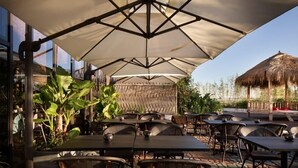 Un área de comedor al aire libre con mesas y sillas bajo un gran paraguas, rodeada de plantas y una estructura de techo de paja.