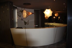 A modern reception desk with a curved design, a wall-mounted butterfly art piece, and pendant lighting. - Ores Boutique Hotel (Bansko)