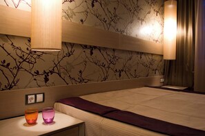 Une chambre d’hôtel moderne avec un grand lit, une table de chevet avec deux verres, un mur orné d’un motif représentant des branches et une lampe suspendue.