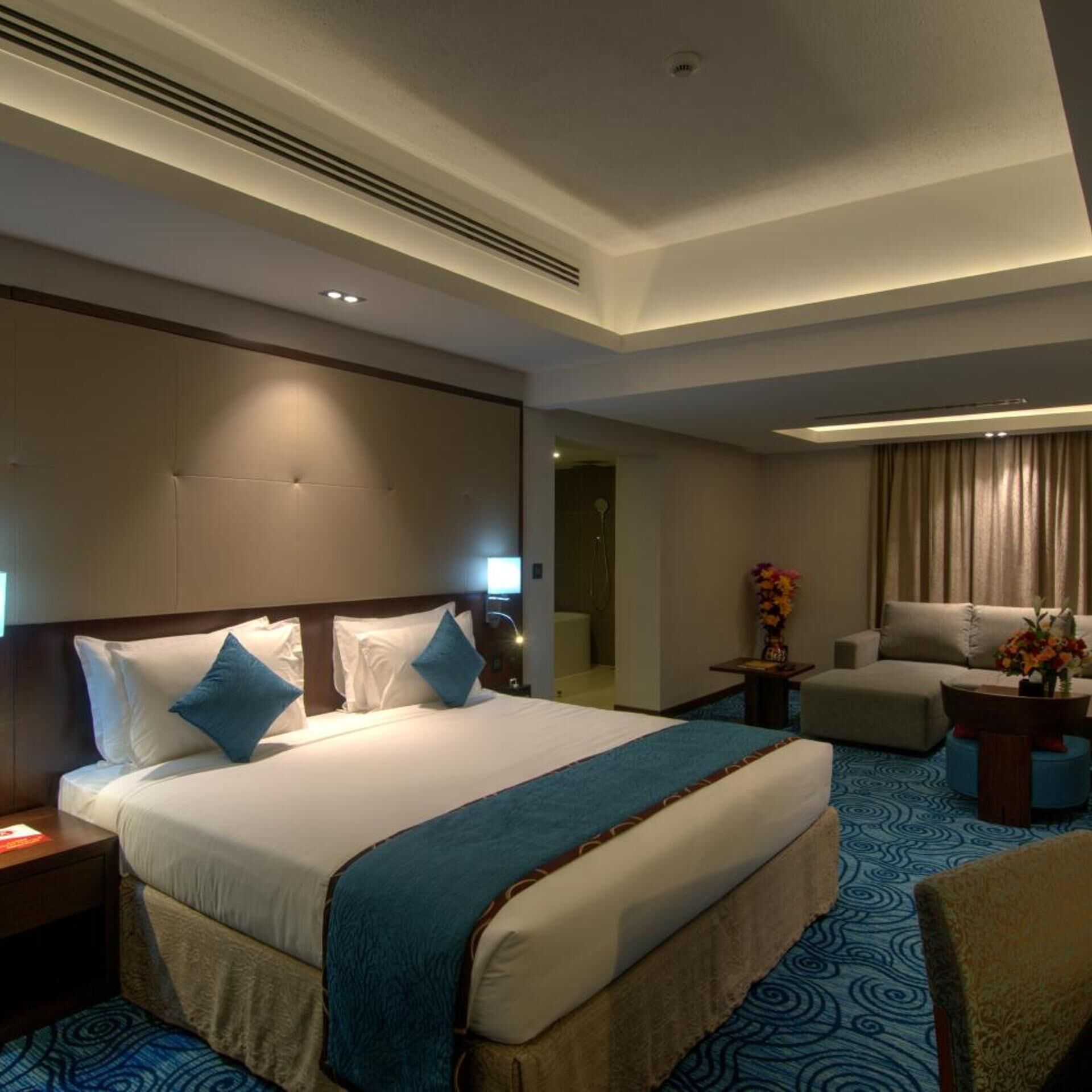 Habitación de hotel con una cama grande, un sofá, una mesita con un jarrón y un baño visible a través de una puerta abierta.