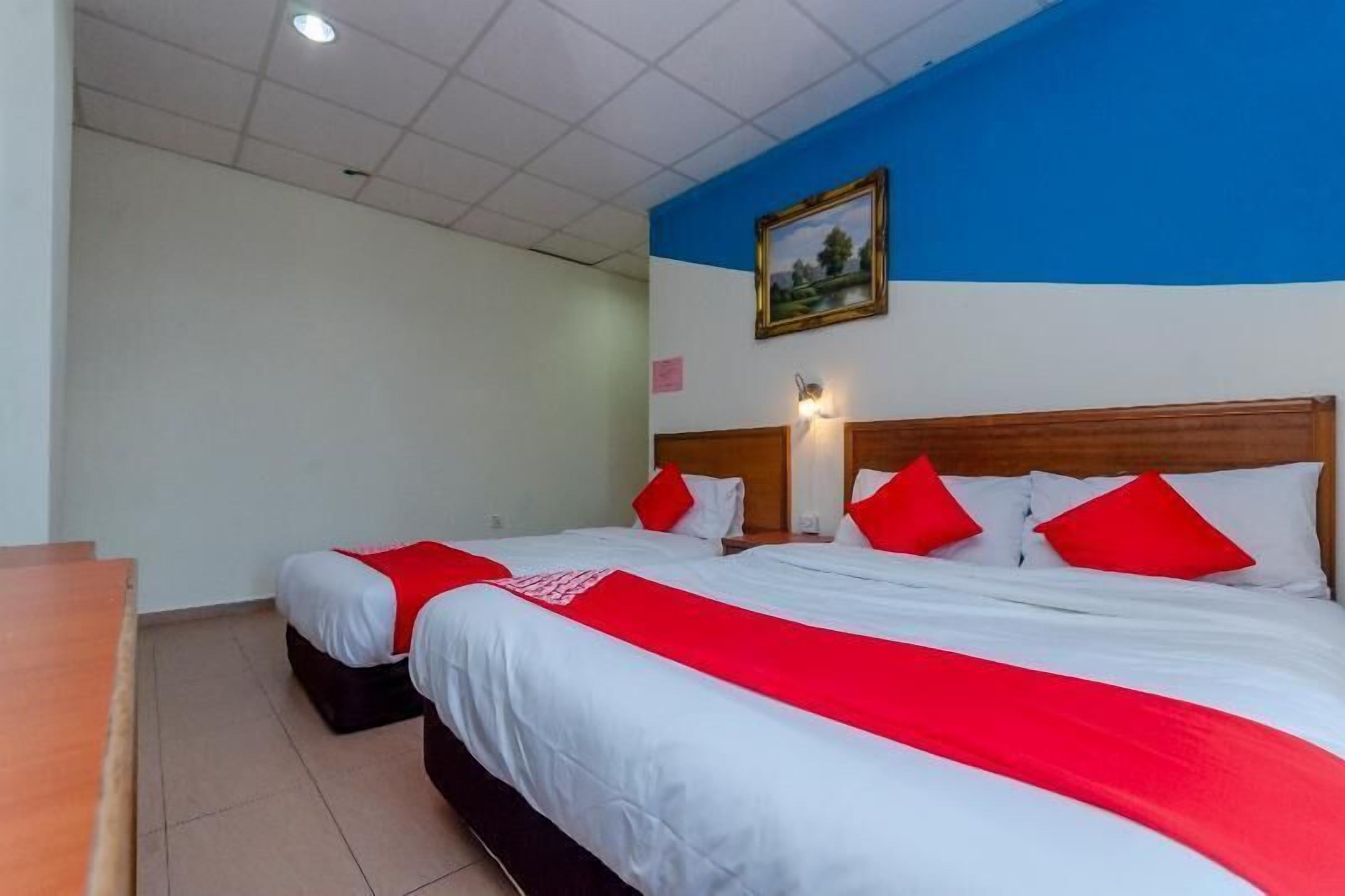 Habitación de hotel con dos camas, cada una con ropa blanca y detalles rojos, cabeceros de madera, un cuadro en la pared y un escritorio con una silla.