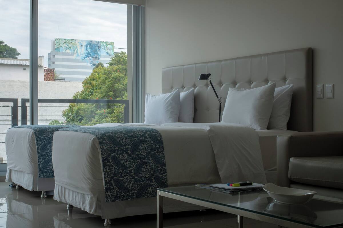 A hotel room with a large bed, a sofa, a glass coffee table, and a view of greenery outside.