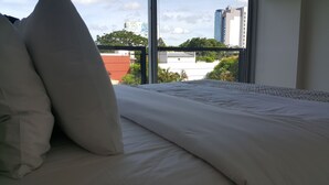 A bed with white bedding and pillows, a window with a view of buildings and trees, and a balcony railing. - Start Villa Morra (Asunción)