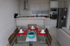 Una cocina moderna con armarios blancos, una campana extractora de acero inoxidable y un comedor con mesa y sillas de vidrio.