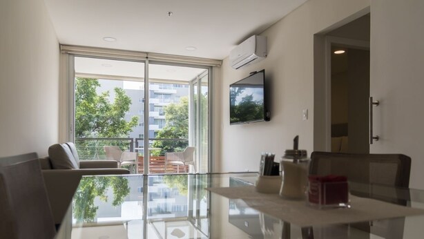 A modern living room with a glass table, a sofa, a TV mounted on the wall, and a balcony with outdoor furniture. - Start Villa Morra (Asunción)