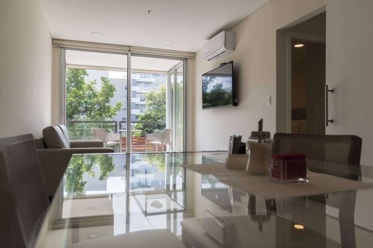 A modern living room with a glass table, a sofa, a TV mounted on the wall, and a balcony with outdoor furniture.