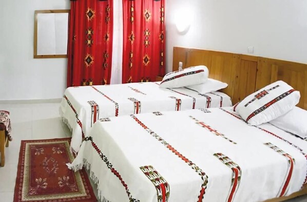 A double bed with white bedding and red and black patterns, a wooden headboard, a red patterned curtain, and a small red rug. - Hotel Louss El Oued (El Oued)