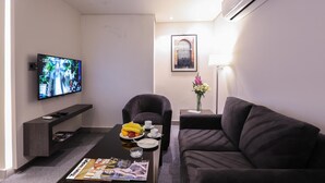 A modern living room with a flat-screen TV, a sofa, a coffee table with fruits and a magazine, and a framed picture on the wall. - Khuttar Furnished Apartments (Amman)