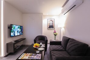 A modern living room with a flat-screen TV, a sofa, a coffee table with fruits and a magazine, and a framed picture on the wall. - Khuttar Furnished Apartments (Amman)