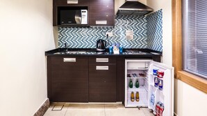 A compact kitchen with dark wooden cabinets, a microwave, a kettle, and a refrigerator stocked with beverages. - Khuttar Furnished Apartments (Amman)