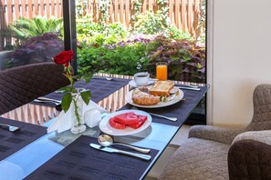 Ett frukostbord dukat med en croissant, vattenmelon, apelsinjuice och kaffe, med en vas med rosor och utsikt över en trädgård genom ett fönster.