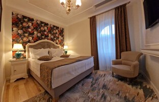 Un dormitorio con una cama grande, dos mesitas de noche con lámparas, una silla, un papel tapiz con estampado floral y un televisor en la pared.