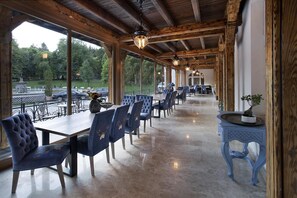 A covered dining area with wooden beams, blue upholstered chairs, and a view of a park. - Hotel Caraiman (Sinaia)