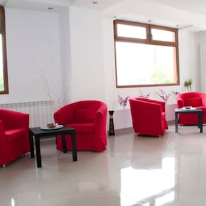 Una habitación moderna con un ventanal grande, sillas rojas, una mesita pequeña y un jarrón con flores.
