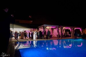 Ett evenemang utomhus som pågår på kvällen, med en pool och ett avskärmat område där folk samlas.