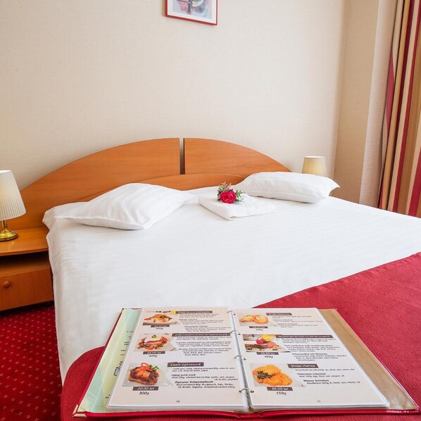Habitación de hotel con cabecera de madera, ropa de cama blanca, manta roja y alfombra roja, además de un menú sobre la cama.