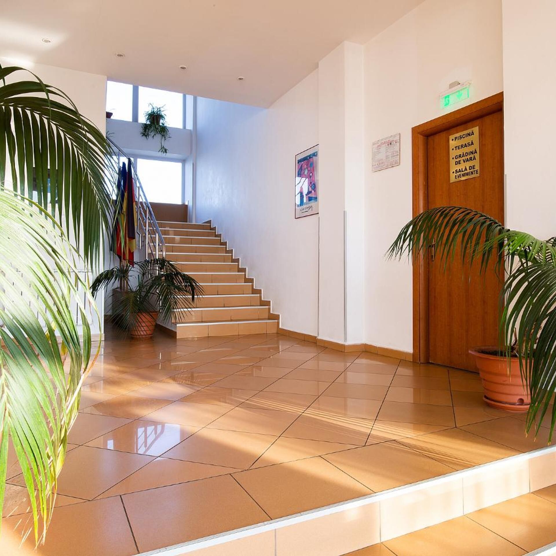 A hotel lobby with a staircase, potted plants, and a wooden door labeled 'PISCINA'.