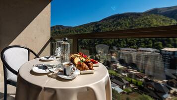Una mesa servida con platos, una copa, una botella y un croissant en un balcón con vistas a un paisaje montañoso.
