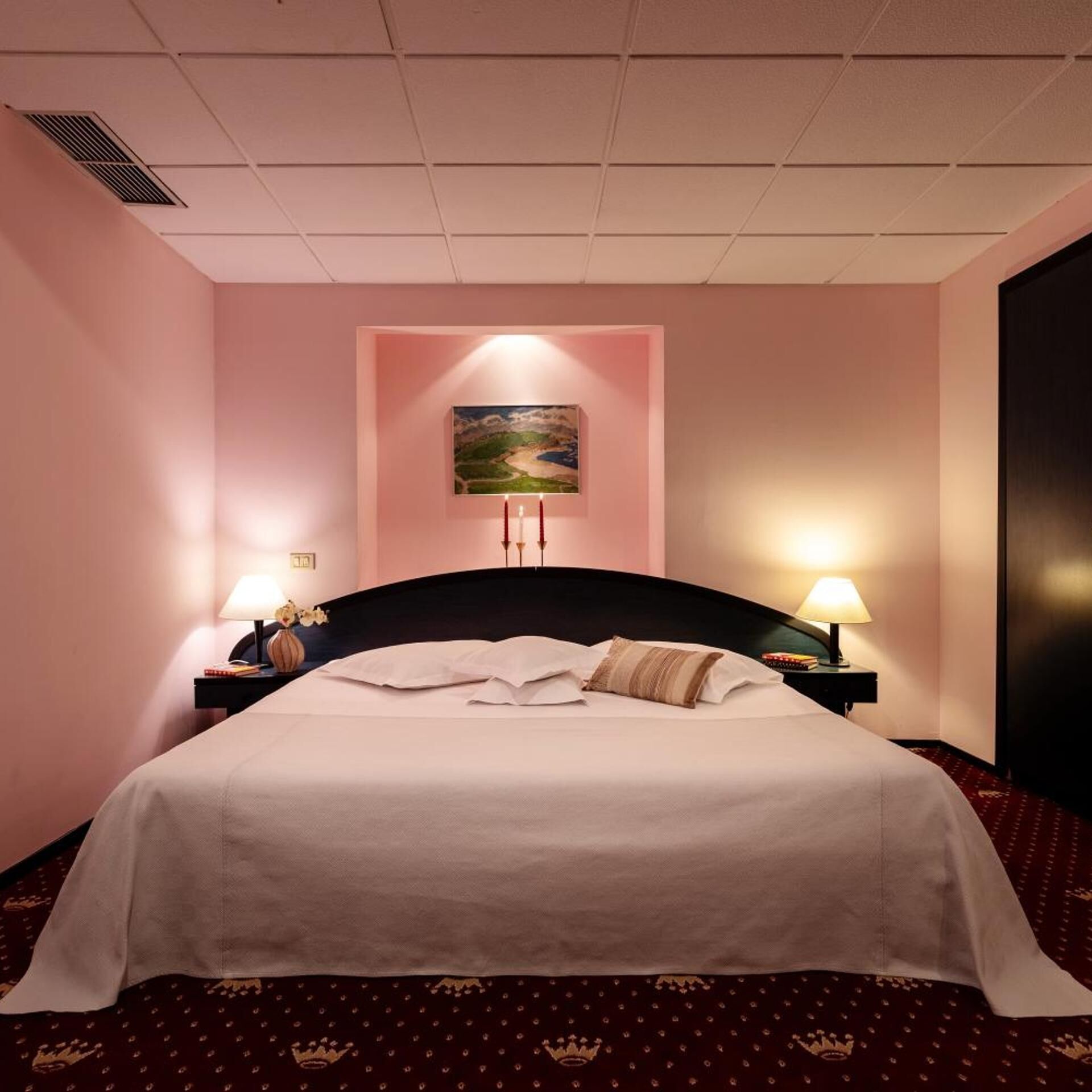 A hotel room with a bed, two bedside lamps, a painting on the wall, and a dark-colored headboard.