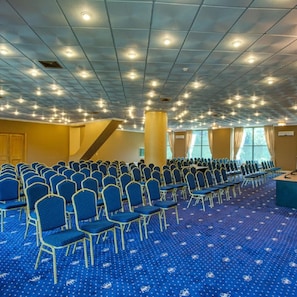 Uma sala de conferências com fileiras de cadeiras azuis, piso coberto de carpete e teto com iluminação embutida.