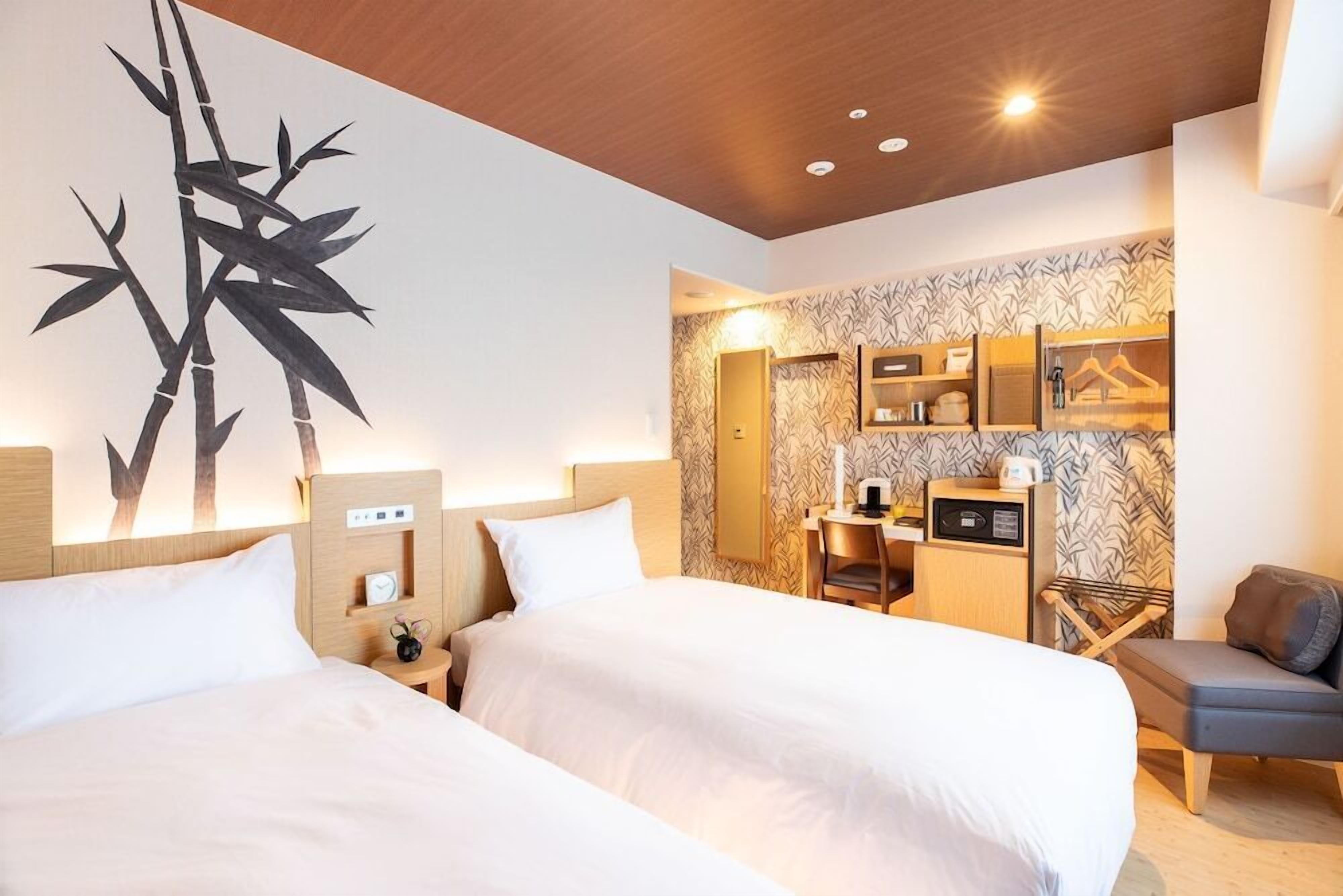A hotel room with two beds, a wall-mounted artwork of bamboo, a desk with a chair, and a small seating area.