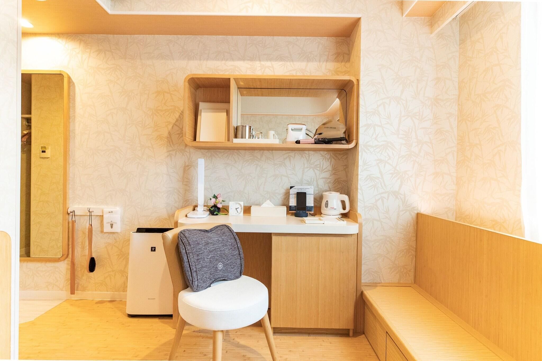 A compact, well-organized room with a small desk, a chair, a compact kitchenette, and a built-in shelf above the desk.